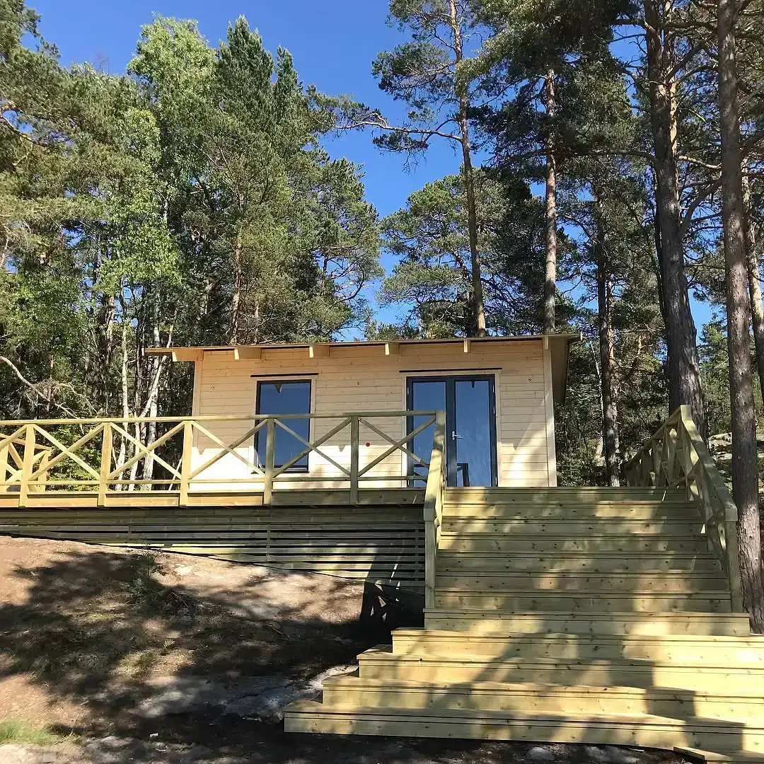 Ljust trähus med altan och trappa i skogsmiljö under blå himmel.