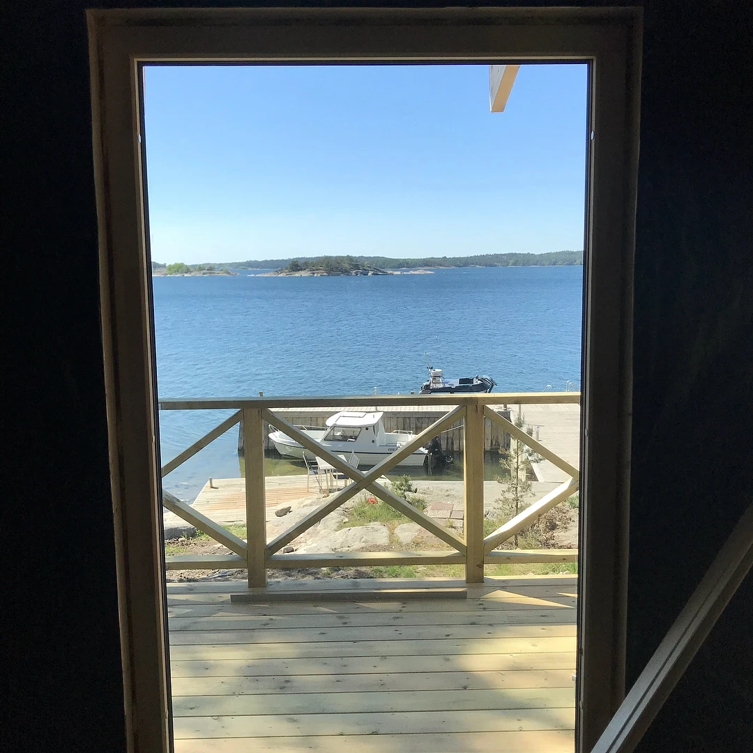 Utsikt genom dörröppning mot hav, brygga, båtar och kust på solig dag.