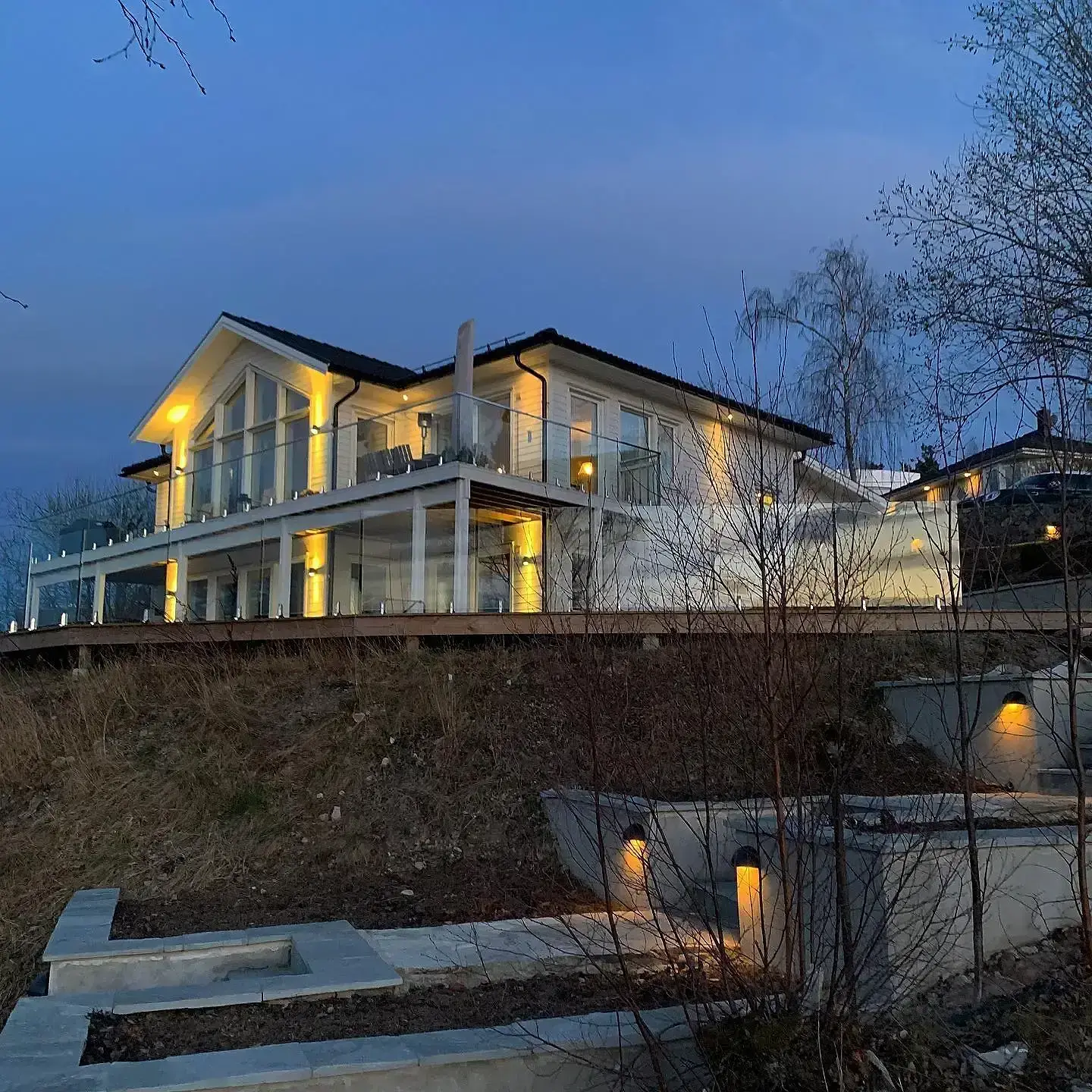 Modern tvåvåningsvilla med upplyst fasad vid skymning på sluttande tomt.